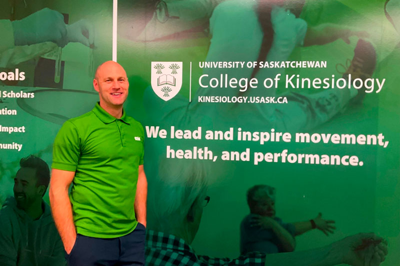 Blair Healey stands in the College of Kinesiology at the University of Saskatchewan in front of a green wall with images of people stretching
