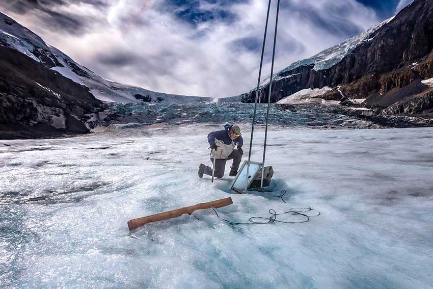 USask researcher, John Pomeroy kneels on the Athabasca Glacier inspecting a monitoring station sitting on the ice