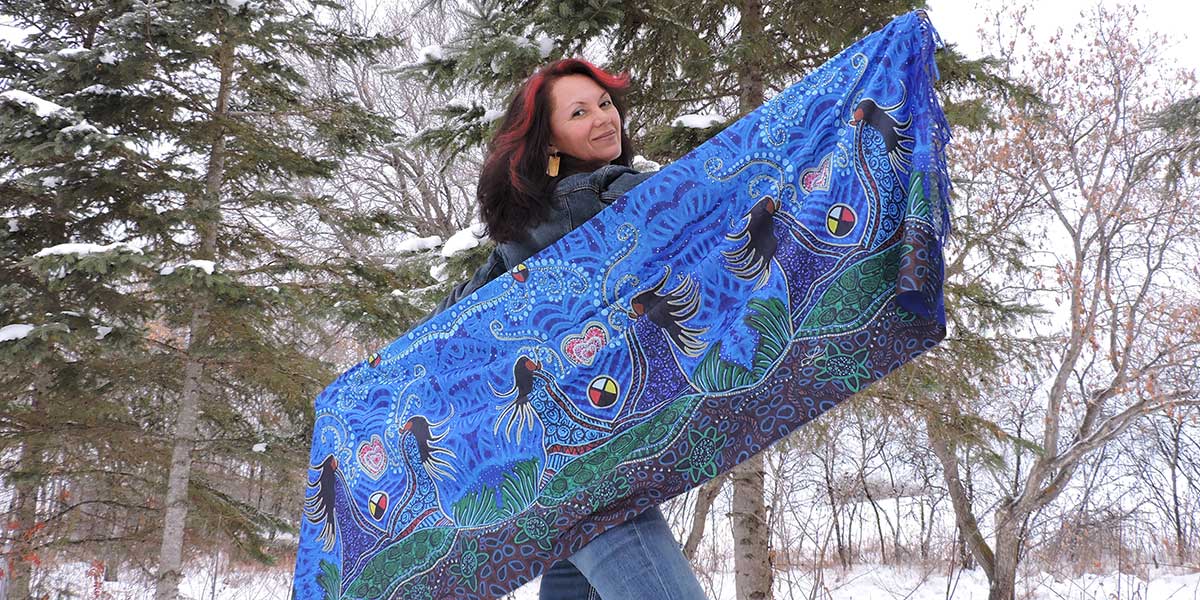 Leah Dorion is standing outside in the snow holding up a colourful blue scarf behind her.